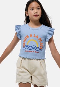 Un crop top côtelé bleu clair avec des manches volantes, mettant en avant un design graphique d'un soleil et des vagues dans des tons orange et bleu. Un short crème avec un ourlet festonné.