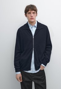 Námořnický zipový svetr přes světle modrou košili, s vysokým límcem, žebrovanými manžetami a měkkou texturou, zkombinovaný s tmavě šedými kalhotami.