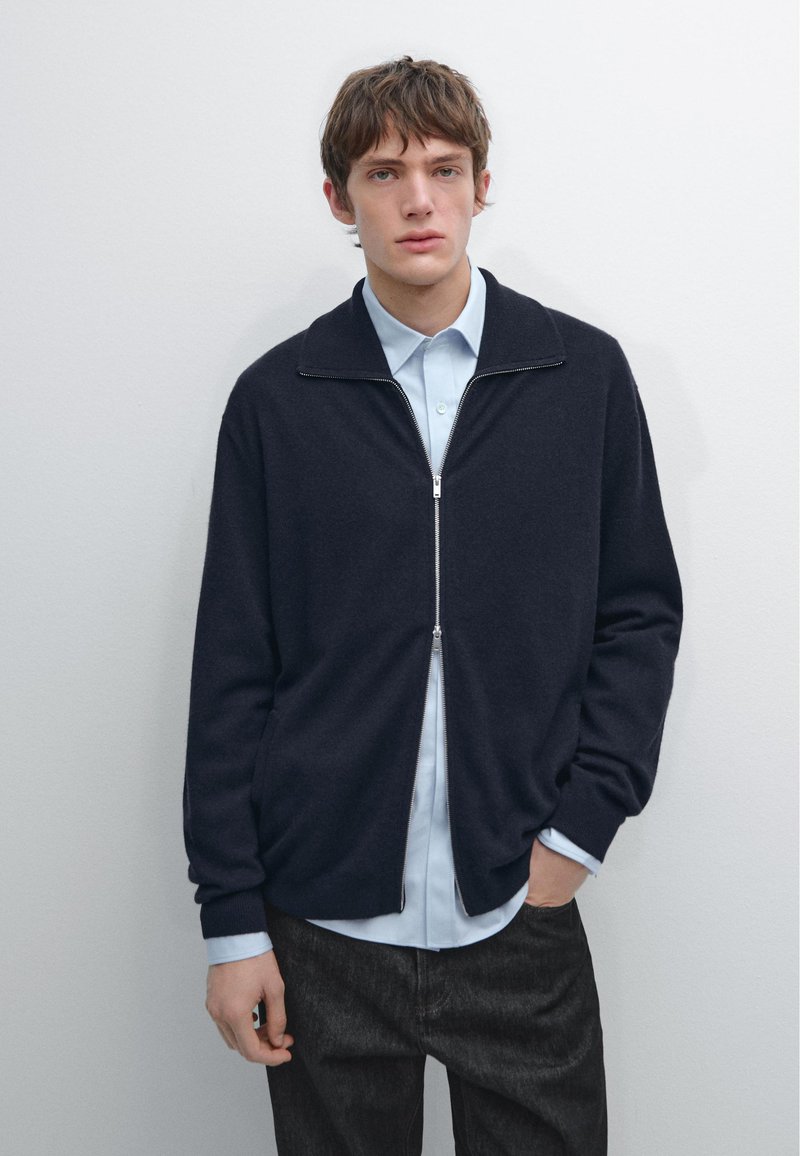 Námořnický zipový svetr přes světle modrou košili, s vysokým límcem, žebrovanými manžetami a měkkou texturou, zkombinovaný s tmavě šedými kalhotami.