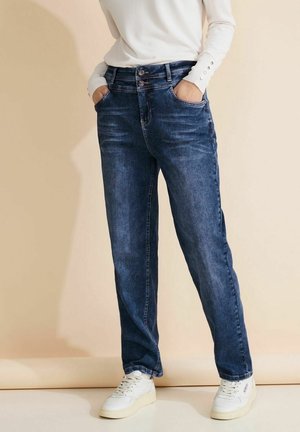 Personne portant un jean bleu à coupe droite, une chemise blanche à manches longues avec boutons aux poignets et des baskets blanches, les mains dans les poches, fond neutre.
