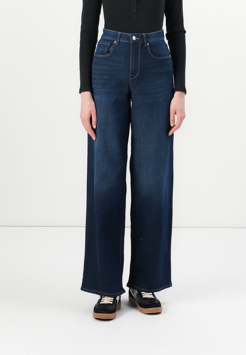 Personne portant un jean taille haute large bleu foncé associé à un haut noir à manches longues et des baskets noires avec des lacets blancs.