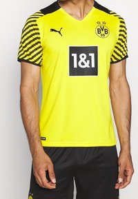 Gul fotbollströja med svarta ränder på ärmarna, med V-ringning, logotyp och sponsortryck på framsidan. Mesh-textur.