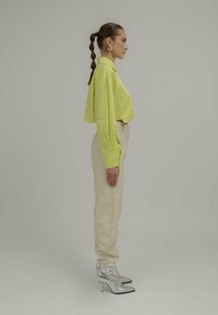 Neon grønn cropped skjorte, kremfargede høyt midje bukser og sølvfargede spisse ankelstøvletter. Modellen står i profil mot en nøytral bakgrunn.