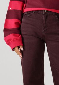 Persoon in hoogwrijdende donker bordeauxrode jeans en een rood met donkerrood gestreepte lange mouwen sweater, met een ontspannen hand naast zich.