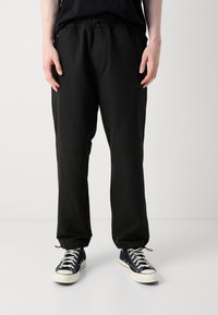 Persoon in zwarte joggerbroek met ritszakken en zwarte high-top sneakers met witte veters en zolen op een effen achtergrond.