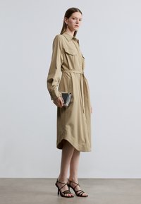 Μπεζ πουκαμισοφόρεμα με μπροστινά κουμπιά, τσέπες στο στήθος, ζώνη στη μέση και μακριά μανίκια με μανσέτες. Συνδυασμένο με μαύρα πέδιλα με λουράκια και ασημένιο τσαντάκι χειρός.