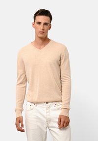 Beige V-hals trui van zacht materiaal; heeft lange mouwen en ribgebreide manchetten. Draagt witte jeans, model staat tegen een neutrale achtergrond.