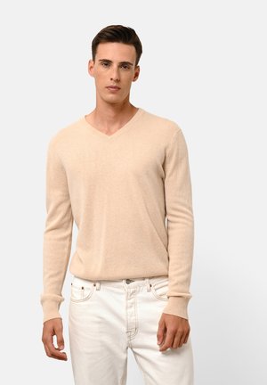 V-neck beige sweater made of soft fabric; features long sleeves and ribbed cuffs. Worn with white jeans, model stands against a neutral background.