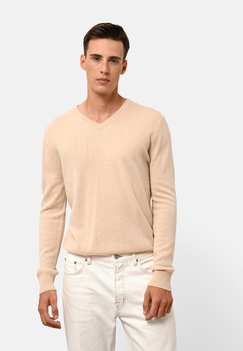 Beige V-hals trui van zacht materiaal; heeft lange mouwen en ribgebreide manchetten. Draagt witte jeans, model staat tegen een neutrale achtergrond.