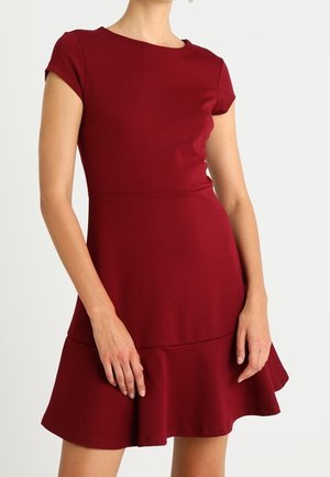 Nainen, joka pukee lyhythihaisen, vartalonmyötäisen viininpunaisen mekon liehukkaan helmaan, seisoo yksinkertaista vaaleaa taustaa vasten.