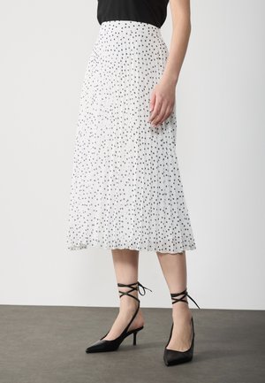 Gonna bianca plissettata con pois neri, caratterizzata da una vita alta e un design fluido. Indossata con tacchi a punta neri con lacci alla caviglia.
