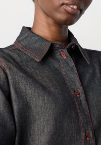 Mørk denimskjorte med kragehals, med røde kontrastsømmer og brune knapper. Nærbilde som fremhever teksturen i stoffet.