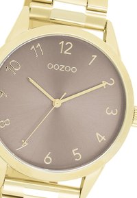 Montre dorée avec un boîtier rond et un cadran taupe mat. Présente des index horaires numériques et des aiguilles fines, attachée à un bracelet métallique.