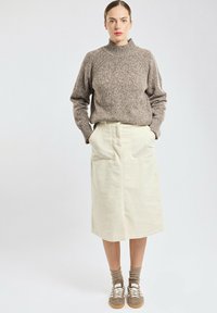 Femme portant un pull en tricot marron, une jupe midi beige avec poches latérales, des chaussettes marron et des baskets beiges, debout les mains dans les poches.