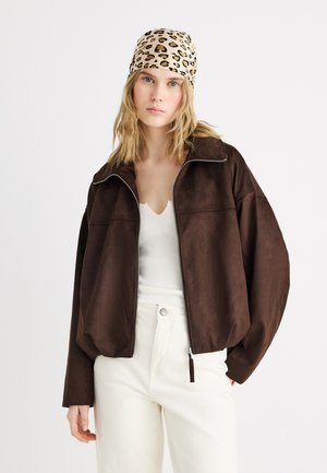 Young woman wearing a leopard-print headscarf, brown zip-up jacket, white ribbed top, and high-waisted white pants standing against a plain background.