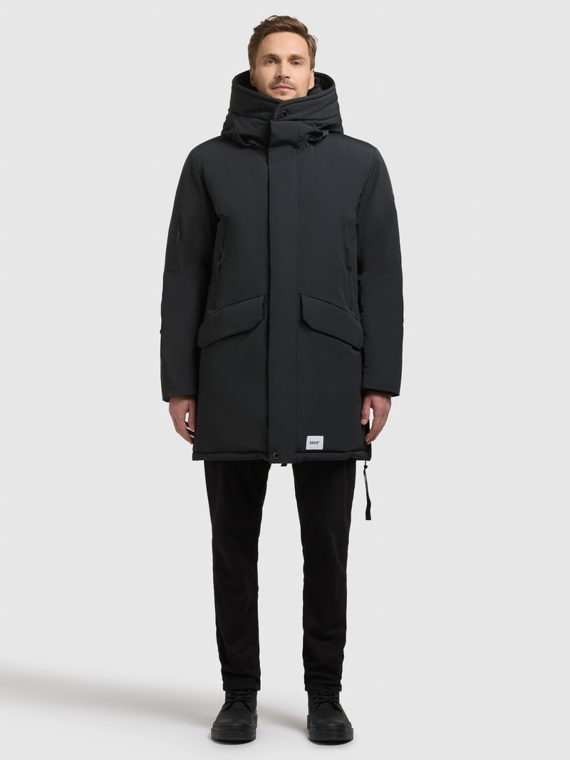 Man standing straight wearing a black hooded winter coat with zip and flap pockets, black pants, and black shoes on a white background.