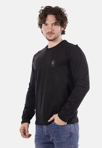 T-shirt nera a maniche lunghe in cotone con colletto rotondo e piccolo logo ricamato sul petto, abbinata a jeans blu.