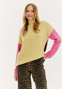 Pletený sveter v žltej farbe s ružovými a hnedými akcentmi, s vysokým golierom a voľným strihom, skombinovaný s nohavicami s leopardím vzorom a malým logom.