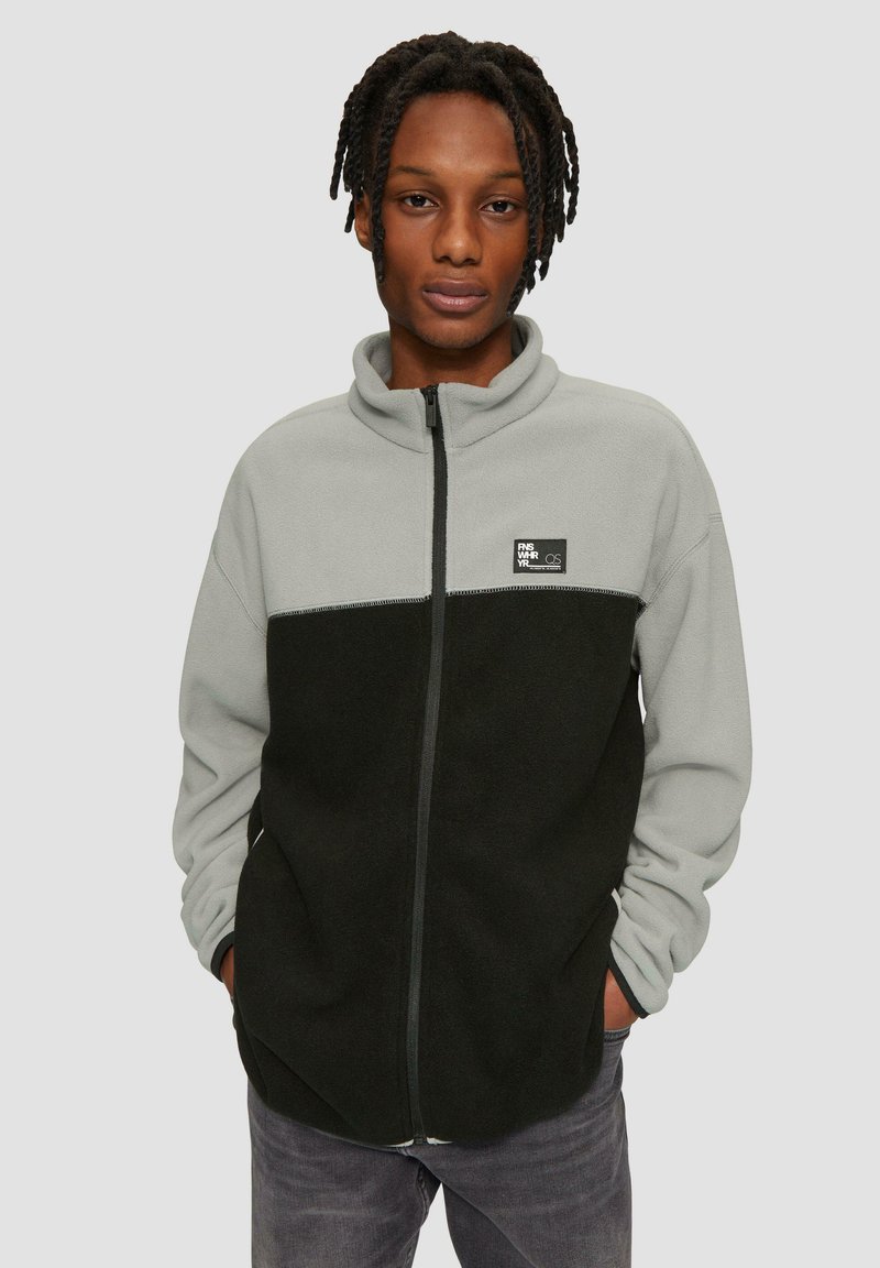 Fleecejack met een grijs bovenste gedeelte en een zwart onderste gedeelte, hoge kraag, volledige rits en een logopatch op de borst. Zachte textuur.