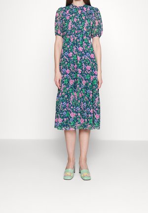 Kobieta ubrana w sukienkę w kwiaty do kolan z krótkimi rękawami w kolorach niebieskim, różowym i zielonym oraz zielone buty na obcasie z pastelowymi klamrami.