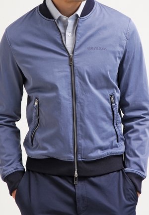 Mann trägt eine hellblaue Armani Jeans Bomberjacke mit Reißverschluss über einem weißen Hemd mit Kragen, die Hände in den Taschen.