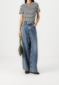 Camiseta a rayas en blanco y negro, vaqueros de mezclilla azul de pierna ancha y botas de tobillo marrones. Bolso verde con herrajes plateados.