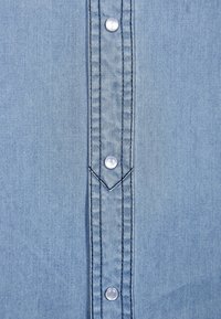 Jasnoniebieska koszula dżinsowa z rzędem pięciu białych guzików, centralnym szwem z zakończeniem w kształcie ostrza oraz subtelną teksturą na całej powierzchni.