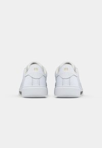 O pereche de adidași albi, prezentată din spate pe un fond alb simplu, având accente cu logo auriu aproape de gulerul călcâiului.