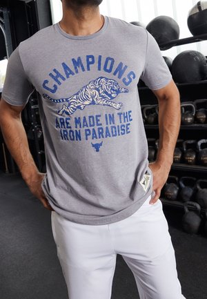Homme portant un t-shirt gris avec texte bleu, graphique de tigre et slogan « Champions are made in the Iron Paradise », debout dans une salle de sport avec des poids.