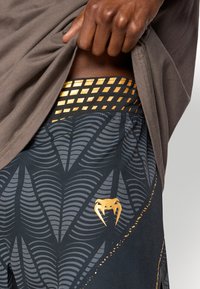 Μαύρα αθλητικά σορτς με υφασμένο σχέδιο φύλλου σε σκούρο γκρι, με χρυσές λεπτομέρειες και σχεδιασμένη ζώνη μέσης. Ένα λογότυπο είναι ορατό  στο κάτω μέρος του ποδιού.