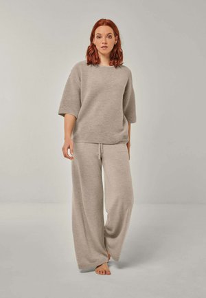 Woman with red hair wearing beige loose knit top and wide-leg pants, standing barefoot against a plain gray background.