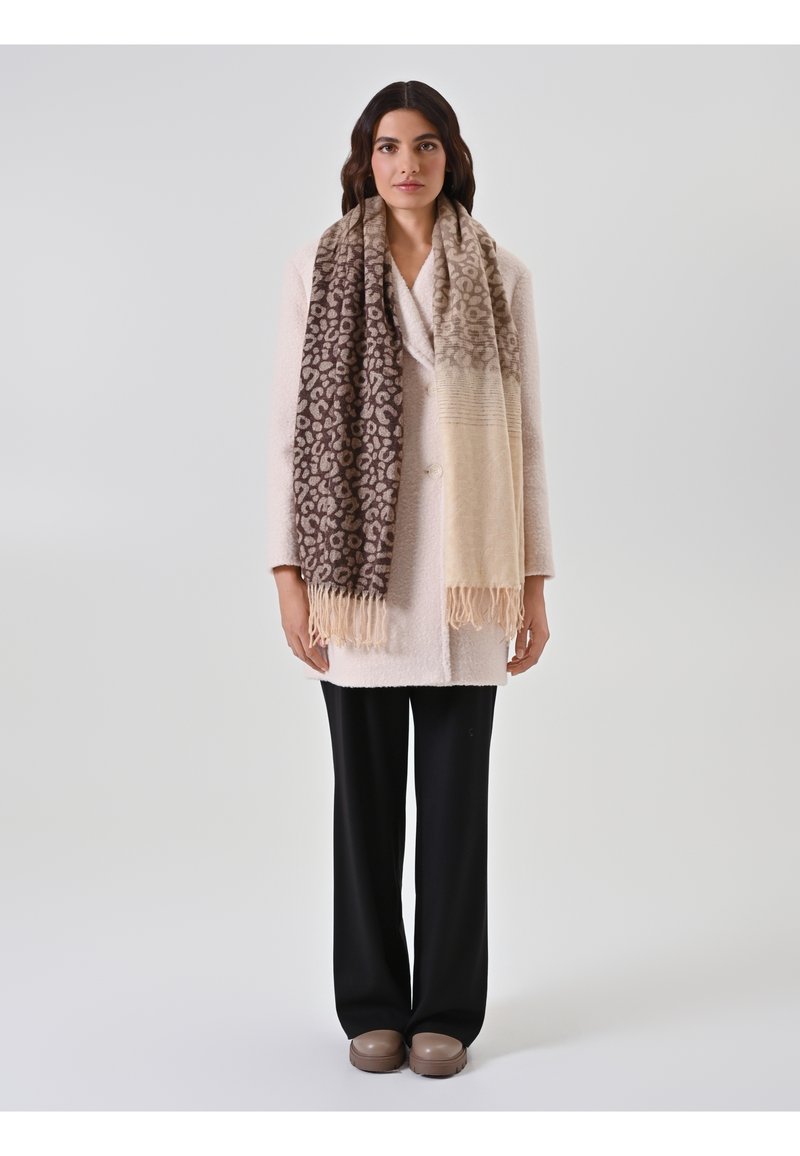 Femme portant un manteau rose clair, une écharpe à imprimé léopard avec des franges, un pantalon large noir, et des chaussures plateformes taupe, se tenant devant un fond uni.