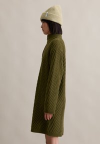 Robe en tricot vert olive avec un col haut, arborant un motif en chevron et des poignets côtelés. Associée à un bonnet beige clair.