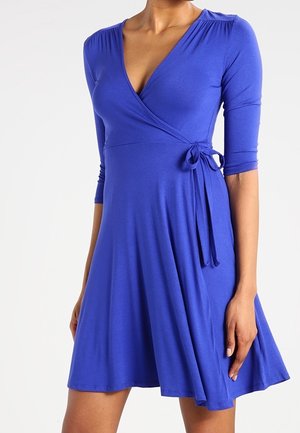 Femme portant une robe portefeuille bleu roi, arrivant au genou, avec des manches trois-quarts et un nœud papillon sur le côté, sur un fond blanc uni.