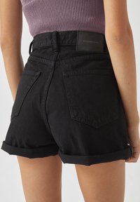 Pantalones cortos de mezclilla negra con dobladillos enrollados, que cuentan con dos bolsillos traseros y un parche de logo negro en la cinturilla. Tejido texturizado y resistente.