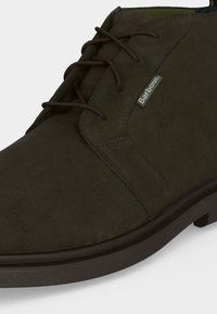 Botte en daim brun foncé à lacets avec des coutures visibles et une petite étiquette logo Barbour en tissu sur le côté près des lacets.