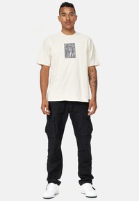 Crème-kleurig t-shirt met een gecropte zwart-wit grafische print in het midden, gecombineerd met zwarte cargobroeken en witte sneakers.