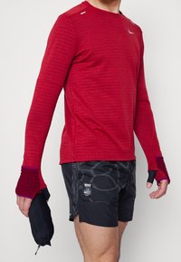 Homme portant un haut de sport rouge à manches longues avec des trous pour les pouces, un short de course noir à motifs, tenant un coupe-vent noir plié dans une main.