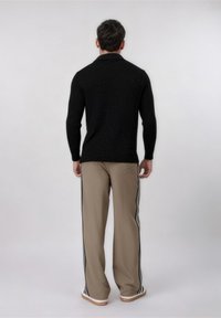 Άνδρας όρθιος με την πλάτη γυρισμένη, φοράει μαύρο πουλόβερ με μακριά μανίκια, μπεζ παντελόνι με μαύρες πλευρικές ρίγες και λευκά αθλητικά παπούτσια με καφέ σόλες.