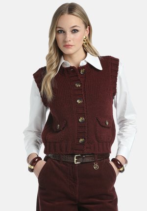 Vīna sarkana adīta veste ar ribotu faktūru, pogām priekšpusē un divām priekšējām kabatām, valkāta pāri baltai kreklam ar apkakli. Bikses un aksesuāri nav attēloti.