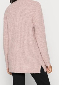 Osoba oblečená v dlouhém, růžovém pleteném svetru s bočními rozparky a černými kalhotami, zobrazená zezadu na čistém pozadí.