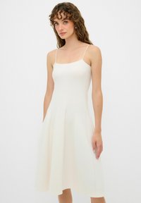 Un vestito senza maniche di un leggero color crema con un corpino aderente e una gonna svasata. Realizzato in un tessuto a costine, presenta sottili spalline a spaghetti.