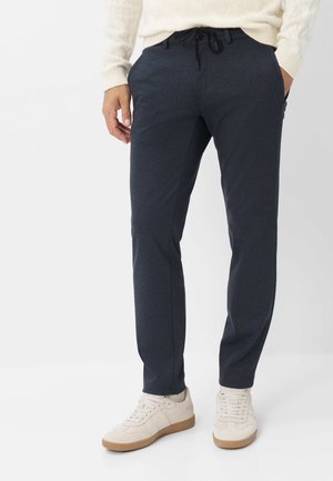 Uomo che indossa pantaloni blu navy con coulisse, maglione crema e scarpe da ginnastica bianche in piedi davanti a uno sfondo uniforme.