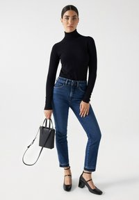 Top ajustado de cuello vuelto negro; jeans azules de cintura alta con dobladillo deshilachado; zapatos negros de tacón cuadrado; pequeño bolso negro con correa para el hombro.