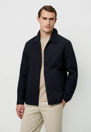 Marineblaue Jacke mit Kragen, vorderen Knöpfen und seitlichen Taschen, getragen über einem hellbeigen Hemd. Das Material wirkt glatt und robust.