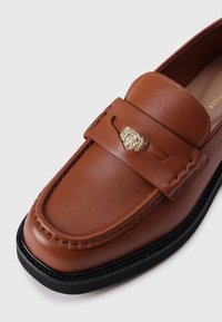 Mocassin en cuir marron avec un emblème doré, des détails de couture, un bout arrondi et une semelle en caoutchouc noir. Texture lisse avec une finition polie.