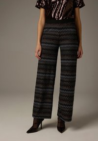 Weit geschnittene Hosen mit einem Zickzackmuster in dunklen Tönen und bordeauxroten Akzenten. Das Material wirkt strukturiert und ist mit einem Paillettenoberteil kombiniert.