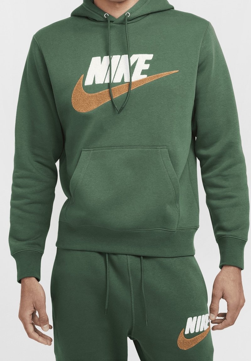 Zelená mikina s klokaní kapsou, s bílým logem Nike a oranžovým znakem swoosh. Materiál vypadá měkký a strukturovaný.