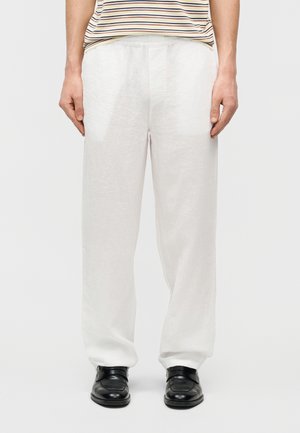 Person iført løstsiddende hvide linnedbukser, sorte lædersko og en indstoppet, kortærmet, stribet skjorte, stående mod en ensfarvet baggrund.