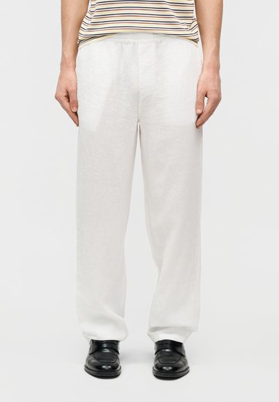 Personne portant un pantalon ample en lin blanc, des chaussures en cuir noir et une chemise à manches courtes rayée rentrée dans le pantalon, debout devant un fond uni.